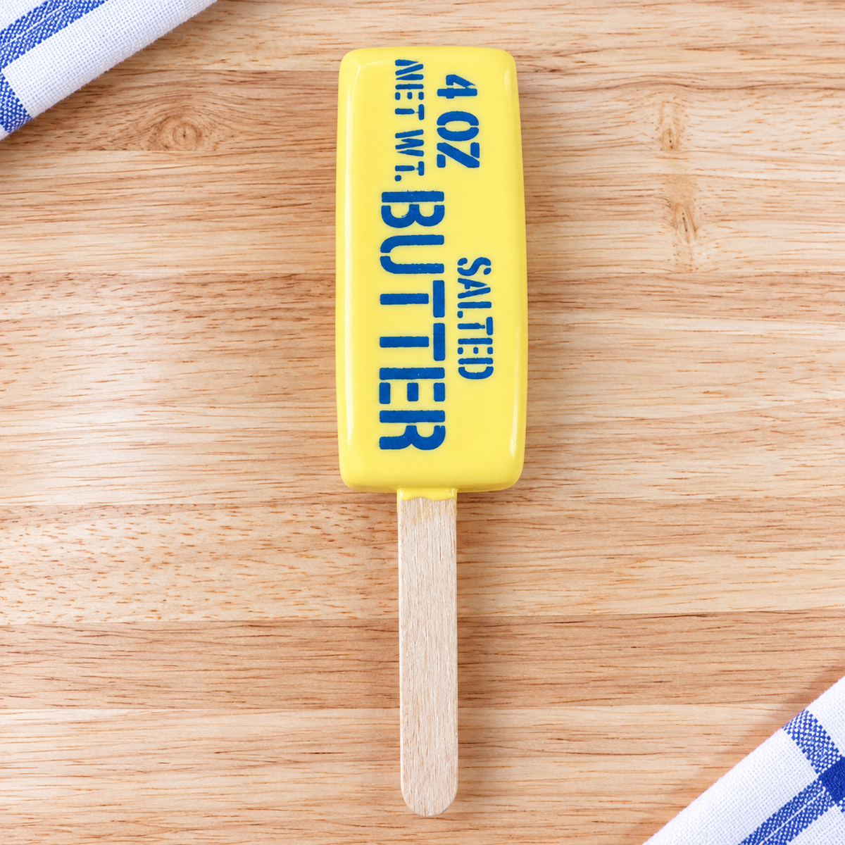 A novelty butter-shaped popsicle with a bright yellow body, blue text reading '4 OZ SALTED BUTTER NET WT.', and a wooden stick, placed on a light wood surface with blue-and-white striped cloth corners visible.
