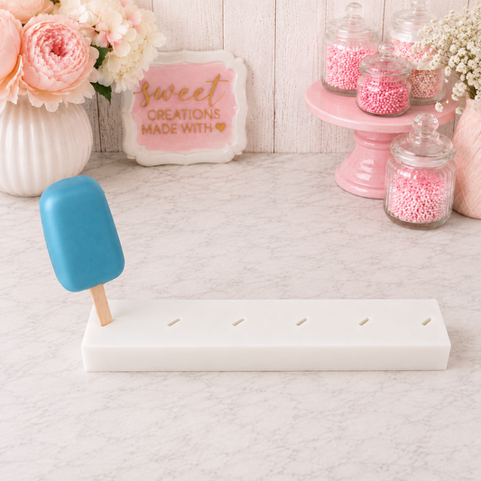A white 6 hole cakesicle stand, with angled slots., holding a blue cakesicle on a wooden stick.