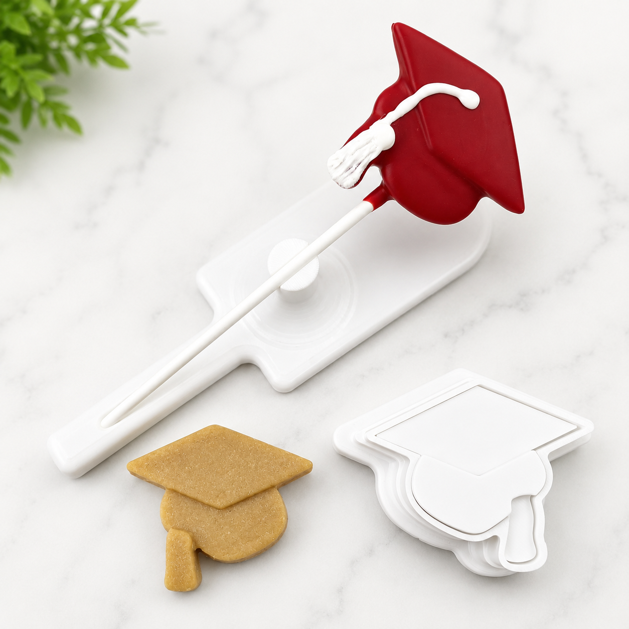 A graduation cap cake pop cutter set on a white marble surface, featuring a white graduation cap cutter, and a cake pop cutout.