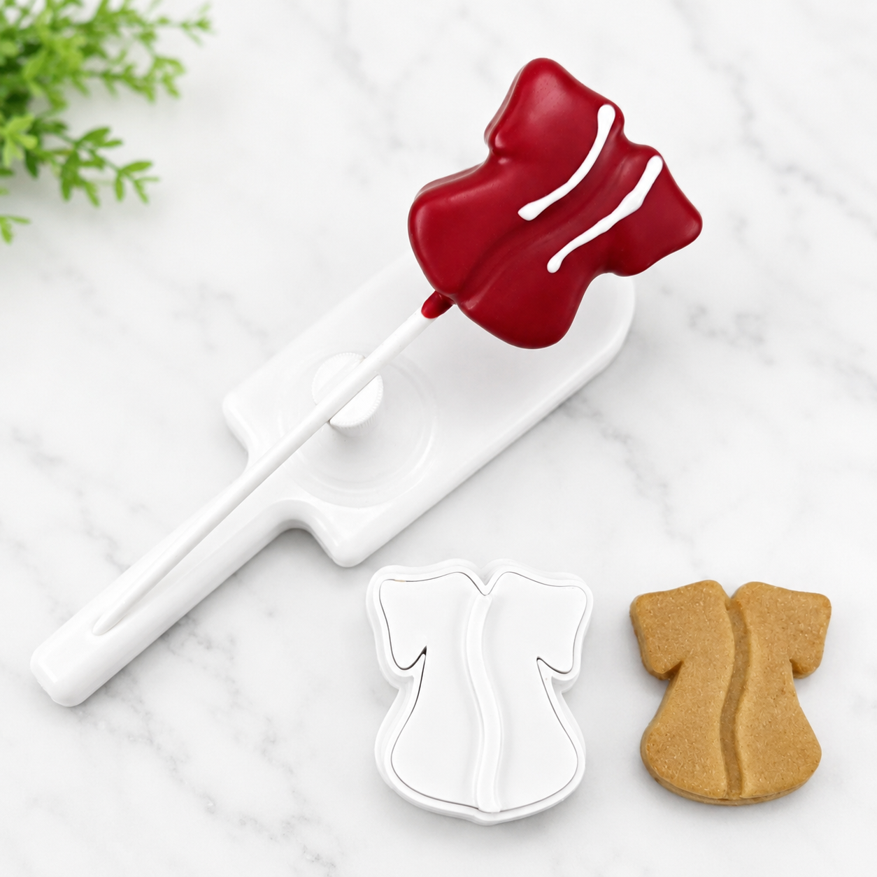 A white plastic cake pop cutter shaped like a Graduation Gown is shown with a red Gown Cake pop  a plain cake pop  cutout on a white marble surface.
