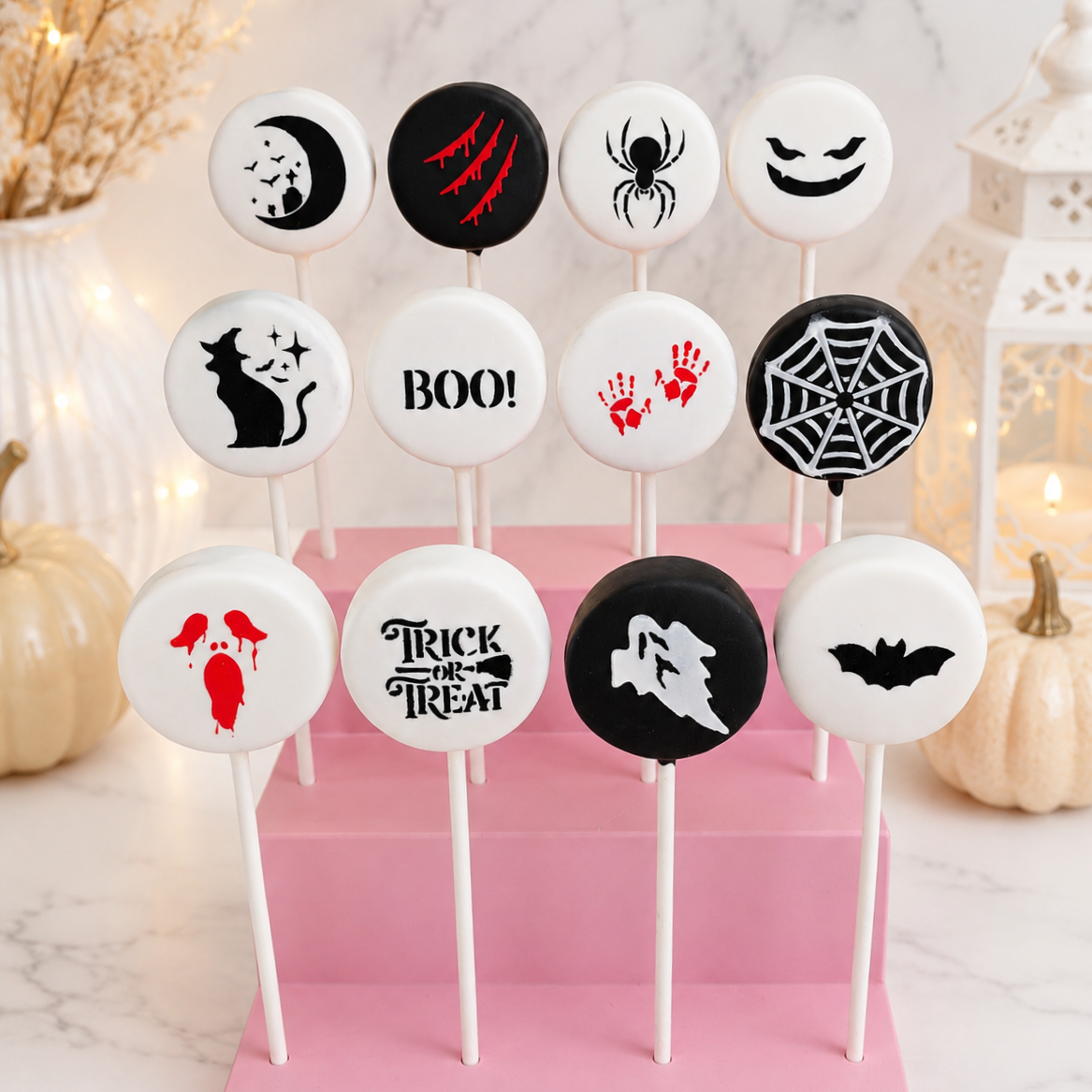Twelve Halloween-themed round cake pops on white sticks, arranged on a pink display block. The designs include a moon and stars, claw marks, a spider, a smiling face, 'BOO!', red handprints, a spiderweb, a cat, a wolf howl, a bat, and a skull with dripping blood motifs.