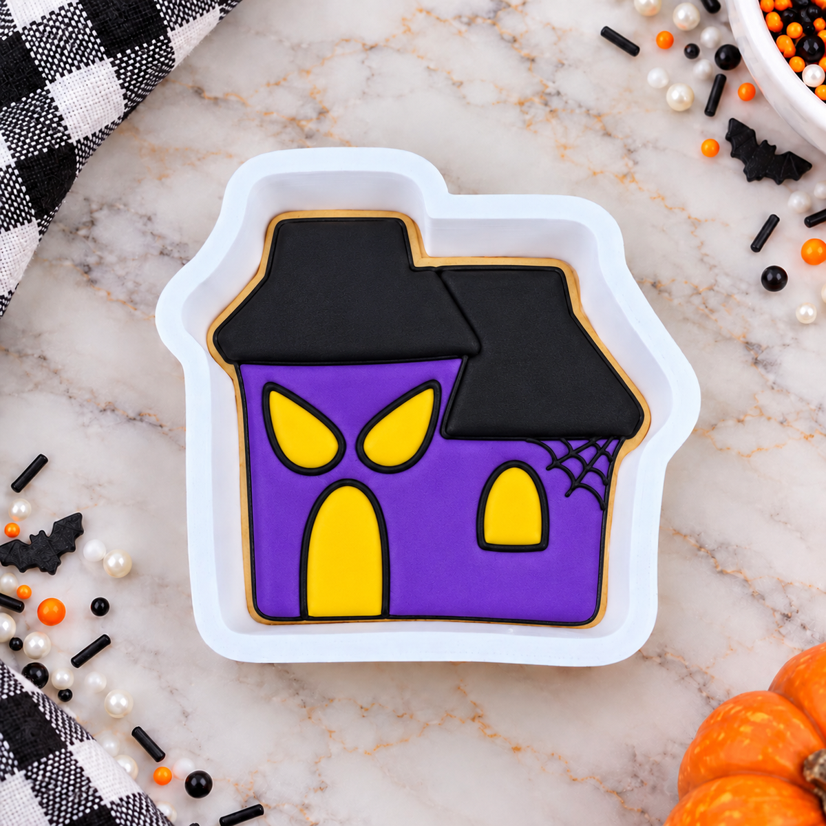 A Halloween haunted house shaped cookie with purple icing, yellow windows and door, and black roof, presented in a white cookie cutter on a marble surface with Halloween decorations.