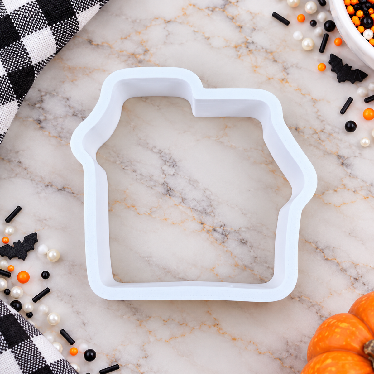 A white house-shaped cookie cutter or mold placed on a white marble surface with Halloween-themed decorations including black-and-white buffalo plaid fabric, candy, sprinkles, and small pumpkins.