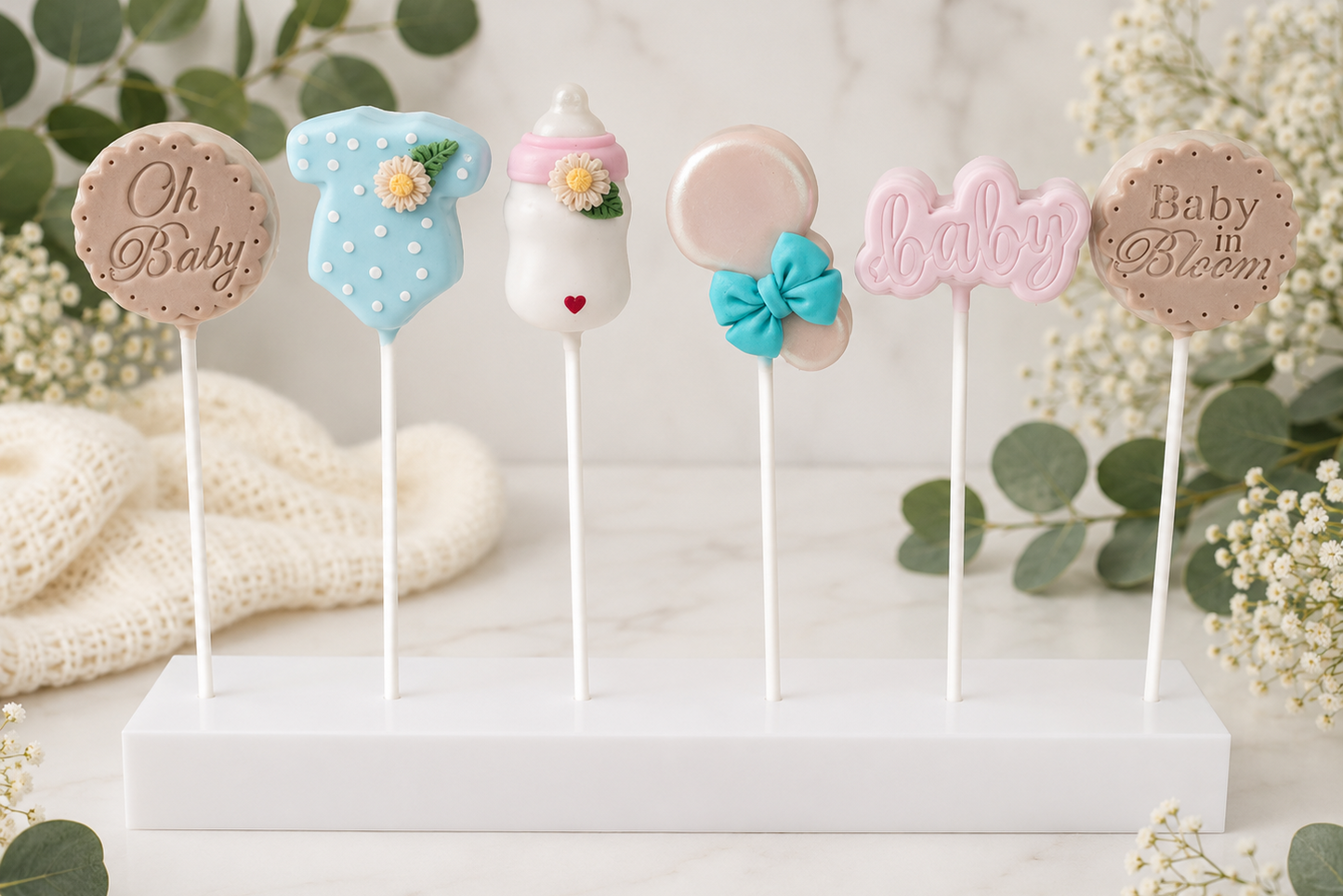 A set of baby-themed cake pops on a white display stand, decorated with fondant shapes including a blue onesie, a baby bottle, a pink balloon, a bow, and the words 'baby' and 'baby in bloom', styled with eucalyptus and baby's breath flowers.